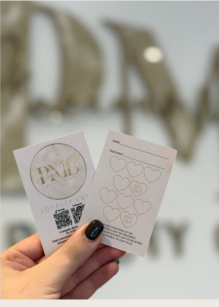 Precious Moments Beauty Clinic loyalty stamp card, front and back, held in front of the PMB wall signage in Banstead