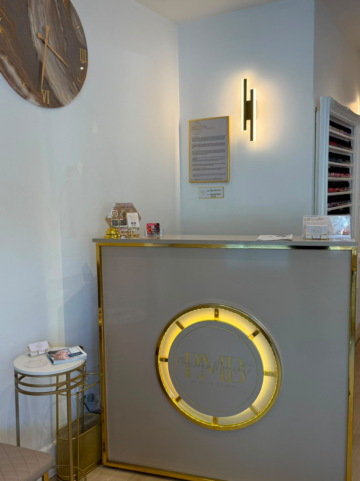 Reception desk with the circular gold PMB logo and marble wall clock at Precious Moments Beauty Clinic, Banstead